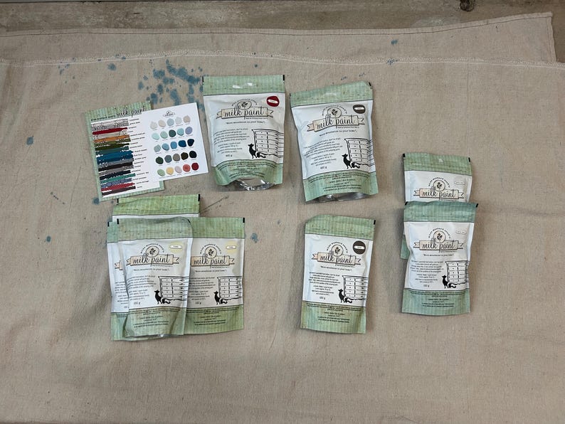 May include: A collection of eight unopened bags of milk paint. The bags are green and white with the words "Milk Paint" printed on them. The bags are arranged in two rows of four.