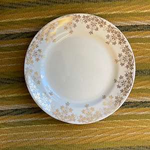 May include: A white ceramic plate with a gold floral design around the rim. The plate is on a striped green and yellow tablecloth.