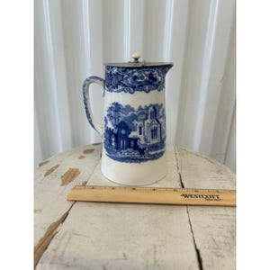 May include: A vintage white and blue ceramic pitcher with a metal lid. The pitcher features a blue transferware design of a church and landscape scene. The handle and rim have blue floral patterns. A ruler is placed next to the pitcher.