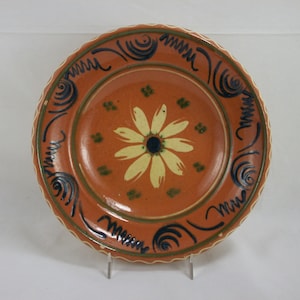 May include: A terracotta plate with a white daisy design and blue and green accents. The plate has a scalloped edge.