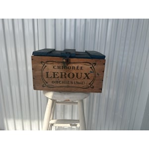 May include: A vintage wooden box with a blue lid and metal latch. The box features the text "CHICORÉE LEROUX ORCHIES (Nord)" in a decorative font. The box is placed on a white stool.