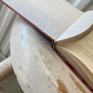 Peut inclure: Un livre reli&eacute; en cuir rouge avec un marque-page en ruban bleu et blanc. Le livre est ouvert &agrave; une page avec du texte manuscrit dans une langue &eacute;trang&egrave;re. Le livre repose sur une surface en bois blanc.