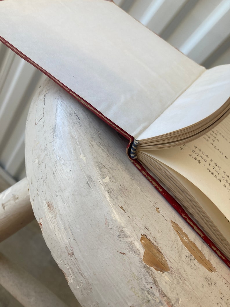Peut inclure: Un livre reli&eacute; en cuir rouge avec un marque-page en ruban bleu et blanc. Le livre est ouvert &agrave; une page avec du texte manuscrit dans une langue &eacute;trang&egrave;re. Le livre repose sur une surface en bois blanc.