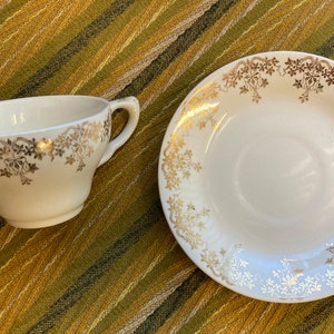 May include: A white teacup and saucer with a gold floral pattern. The teacup and saucer are on a striped green and brown surface.