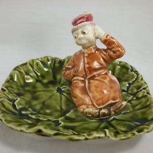 May include: A ceramic figurine of a man wearing a hat and brown jacket sitting on a green leaf-shaped dish.