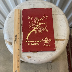 Peut inclure: Un livre rouge avec des lettres dor&eacute;es et une illustration dor&eacute;e d'une branche de cerisier en fleurs. Le titre du livre est "Household Hints in Han Gul".