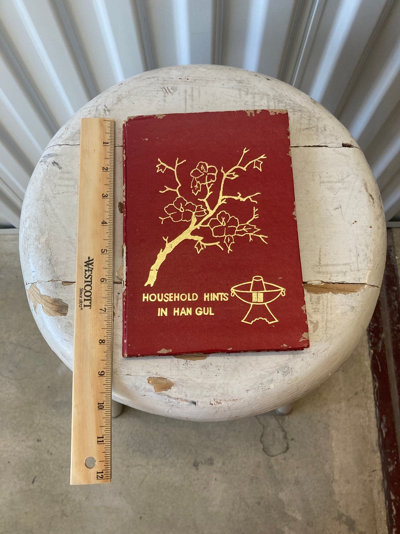 Peut inclure: Un livre rouge avec des lettres dor&eacute;es et une illustration dor&eacute;e d'une branche de cerisier en fleurs. Le titre du livre est "Household Hints in Han Gul".