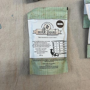 May include: A 230 gram bag of Mustard Seed's Milk Paint in the color "Marigold". The bag is white with green and brown accents. The text "Move mountains in your home." is printed on the bag.
