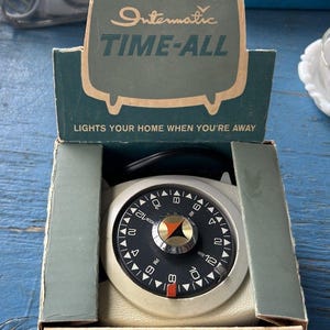 May include: A vintage Intermatic TIME-ALL timer in its original box. The timer features a black and white dial with numbers and a red indicator. The box is teal with the product name and the tagline "LIGHTS YOUR HOME WHEN YOU'RE AWAY".