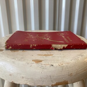 Peut inclure: Un livre reli&eacute; rouge avec une inscription dor&eacute;e sur le dos qui dit "The Year in My Garden". Le livre est pos&eacute; sur un tabouret blanc.