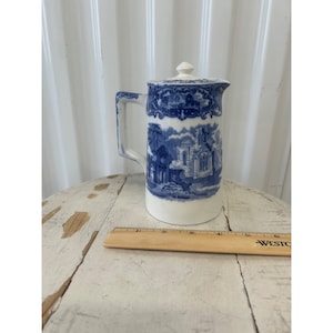 May include: A white and blue ceramic pitcher with a lid. The pitcher features a blue landscape design and a handle. The pitcher is approximately 8 inches tall.