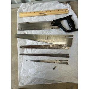 May include: A collection of vintage hand saws and saw blades, including a large saw with a black handle and a wooden ruler with "WESTCOTT" printed on it. The saws are made of metal and show signs of age and use.
