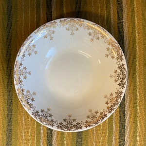 May include: A white ceramic bowl with a gold floral design around the rim. The bowl is empty and sitting on a striped green and brown surface.