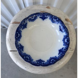 May include: White ceramic bowl with a scalloped edge and a blue floral design. The bowl has a wide rim with a detailed pattern. The bowl is placed on a white, round, distressed wooden surface.