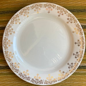 May include: White ceramic plate with a gold floral pattern around the rim. The design features stylized flowers and leaves in a repeating pattern. The plate is round and has a slightly raised edge. The plate is on a green and brown striped surface.