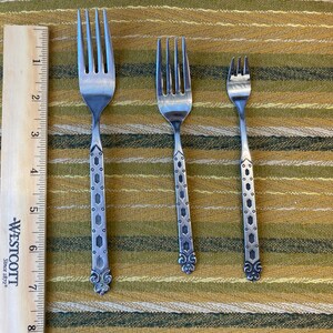 May include: Three silver forks of varying sizes with decorative handles. The handles feature a pattern of small circles and diamond shapes, with a floral design at the base. A wooden ruler is positioned to the left of the forks.