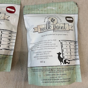 May include: A bag of Mustard Seed's Milk Paint in the color "Marigold". The bag is white with a green and white pattern. The bag has a label with the company logo and the text "Move mountains in your home."