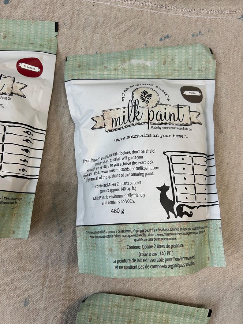 May include: A bag of Mustard Seed's Milk Paint in the color "Marigold". The bag is white with a green and white pattern. The bag has a label with the company logo and the text "Move mountains in your home."