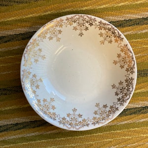 May include: A white ceramic bowl with a gold floral design around the rim. The bowl is on a striped green and yellow surface.