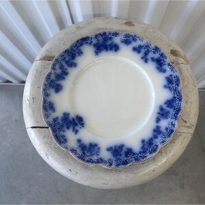 May include: A white plate with a blue floral design around the rim and a gold trim. The plate is placed on a weathered, round, white surface. The plate has a scalloped edge.