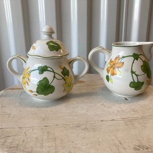 May include: A white ceramic sugar bowl and creamer set with a floral design. The design features yellow flowers and green leaves on a white background.