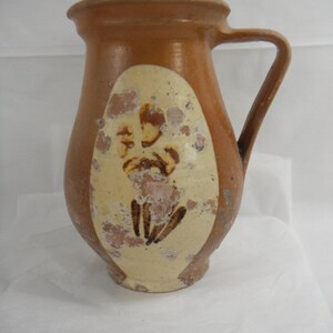 May include: A brown ceramic pitcher with a yellow glaze and a brown design. The pitcher has a handle and a rounded body.