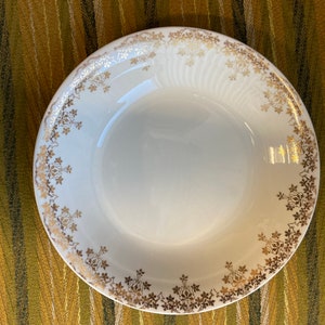 May include: A white ceramic bowl with a gold floral pattern around the rim. The bowl is on a striped green and gold fabric.