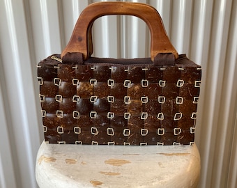 Pode incluir: Uma bolsa de mão marrom tecida com uma alça de madeira. A bolsa é feita de um material tecido que se assemelha a conchas de coco.