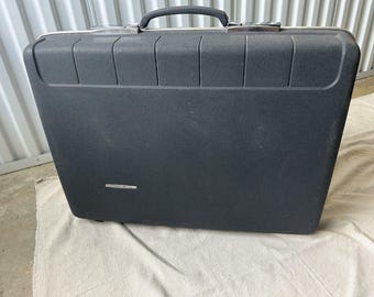 May include: A black vintage suitcase with a textured surface and a silver handle. The suitcase has a rectangular shape with a handle on top and a small silver label on the front. The suitcase is sitting on a white cloth.