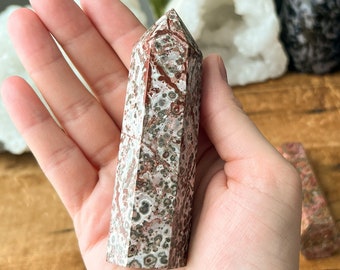 May include: A red and white speckled crystal point, also known as a red jasper point, is held in a hand. The crystal is about 10 cm long and has a pointed tip. The crystal is surrounded by other crystals, including a white crystal cluster and a purple amethyst geode.