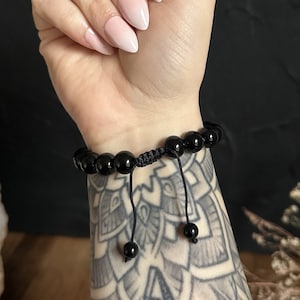 Könnte beinhalten: Schwarzes Perlenarmband mit schwarzem Band und verstellbarem Knotenverschluss. Das Armband wird am Handgelenk getragen.