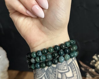 May include: Three green moss agate beaded bracelets are stacked on a wrist with a tattoo. The bracelets are made of small, round beads with a mottled green pattern. The wrist is held up, showing the bracelets and the tattoo.