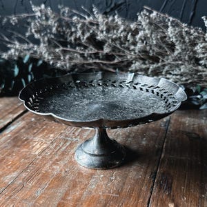 May include: A silver-toned metal pedestal dish featuring a scalloped edge and a decorative pierced border. The circular tray has an intricate engraved pattern on its surface and is supported by a central, flared pedestal base.