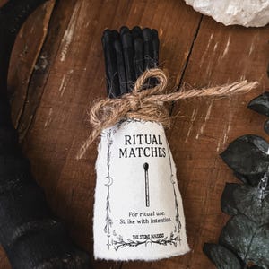 Puede incluir: Un paquete de cerillas rituales con punta negra atadas con hilo. La etiqueta dice "RITUAL MATCHES" con el texto "For ritual use. Strike with intention." Las cerillas están sobre una superficie de madera con un cuerno negro y un cristal en el fondo.