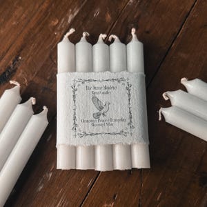 Peut inclure: Plusieurs bougies rituelles blanches, certaines en paquet et d'autres en vrac, disposées sur une surface en bois. Les bougies sont cylindriques avec une finition texturée. Une étiquette avec du texte et un graphisme de colombe entoure un paquet de bougies.