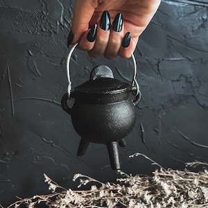 May include: A small black cauldron with a silver handle and three legs. The cauldron is being held by a hand with black nail polish.