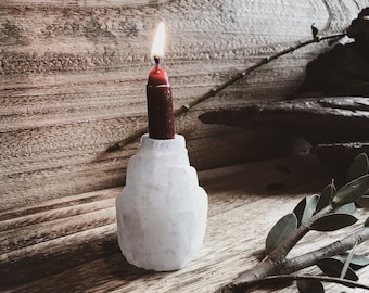 May include: A white selenite candle holder with a red candle burning inside. The candle holder is on a wooden surface with a branch of green leaves.