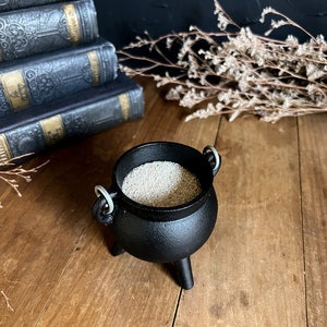 May include: A small black cauldron with three legs and silver handles, filled with white sand. The cauldron is sitting on a wooden surface.