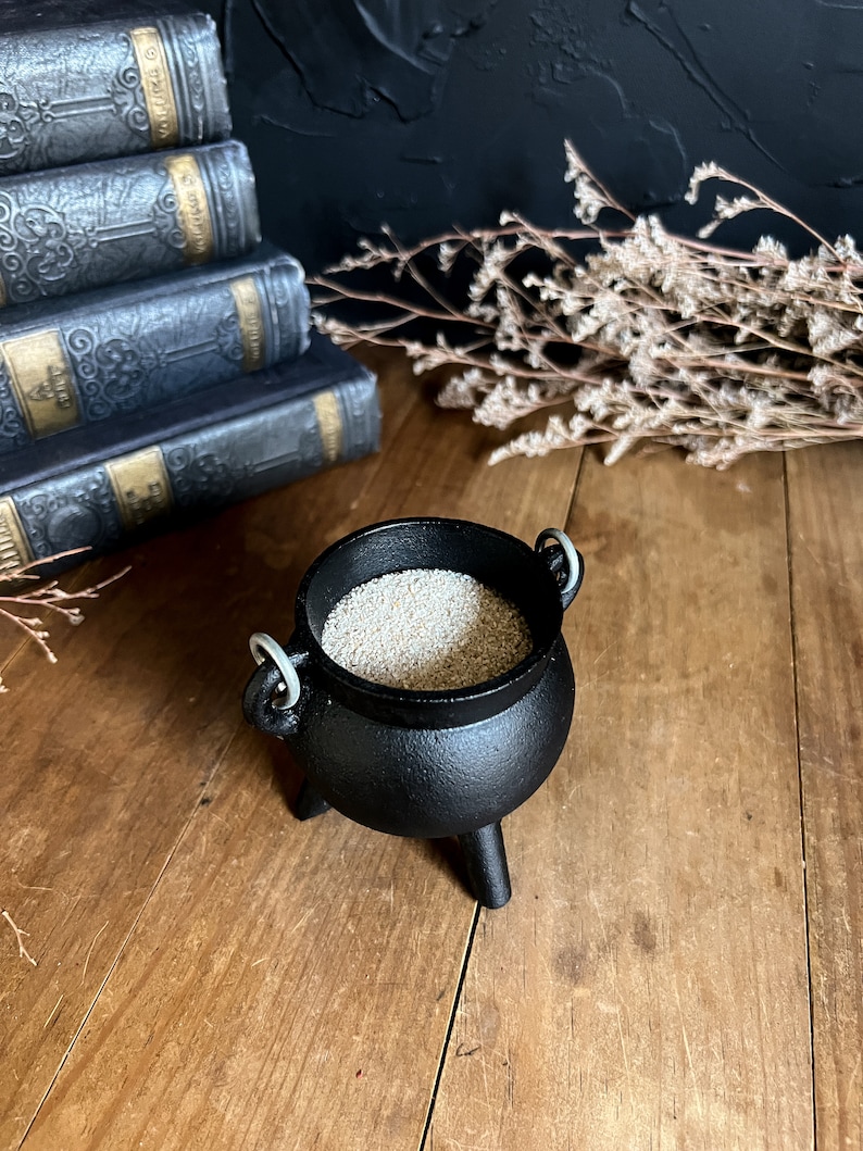 May include: A small black cauldron with three legs and silver handles, filled with white sand. The cauldron is sitting on a wooden surface.