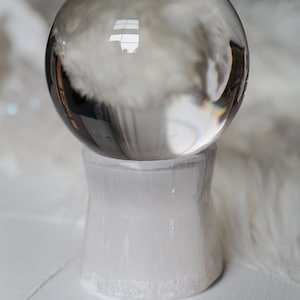Puede incluir: Una bola de cristal transparente descansa sobre un pedestal de selenita blanca. La esfera refleja el entorno, mientras que el pedestal tiene una apariencia texturizada y opaca. La est&eacute;tica general es minimalista y espiritual.