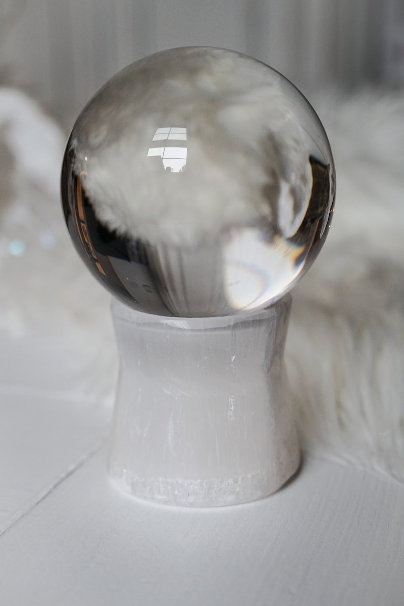 Puede incluir: Una bola de cristal transparente descansa sobre un pedestal de selenita blanca. La esfera refleja el entorno, mientras que el pedestal tiene una apariencia texturizada y opaca. La est&eacute;tica general es minimalista y espiritual.