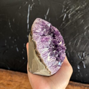 May include: A raw amethyst geode featuring deep purple, crystalline structures within a jagged, hollowed-out stone formation. The exterior of the geode displays a smooth, light brown, natural rock surface, contrasting with the vibrant, multifaceted violet interior crystals.