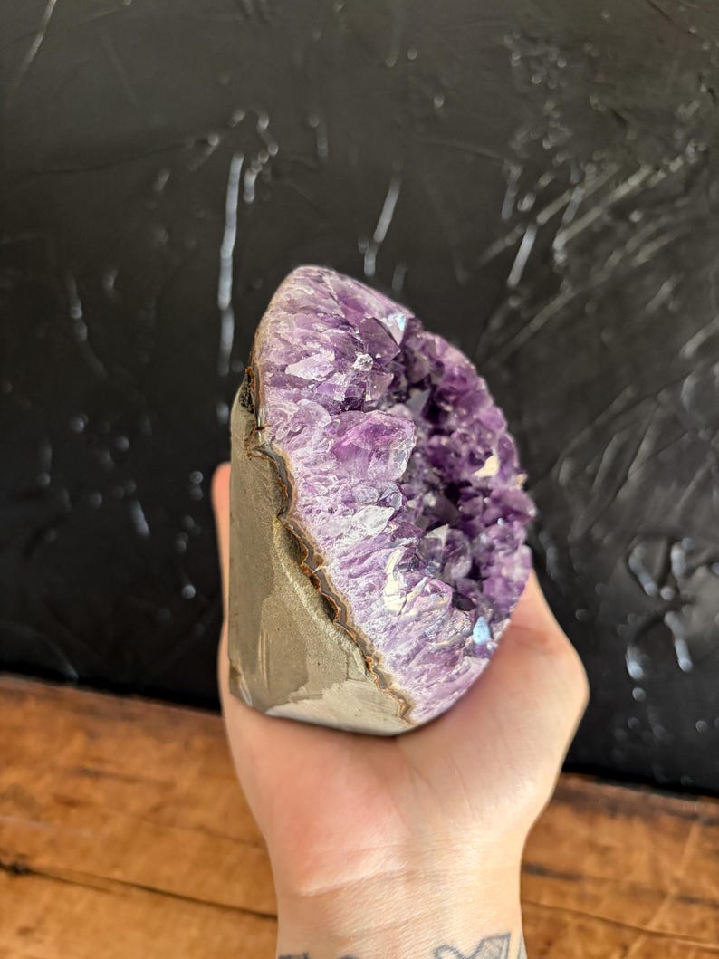 May include: A raw amethyst geode featuring deep purple, crystalline structures within a jagged, hollowed-out stone formation. The exterior of the geode displays a smooth, light brown, natural rock surface, contrasting with the vibrant, multifaceted violet interior crystals.
