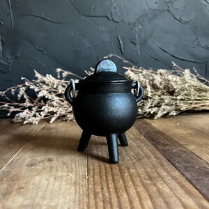 May include: A small black cast iron cauldron with a lid and three legs. The cauldron is sitting on a wooden surface.