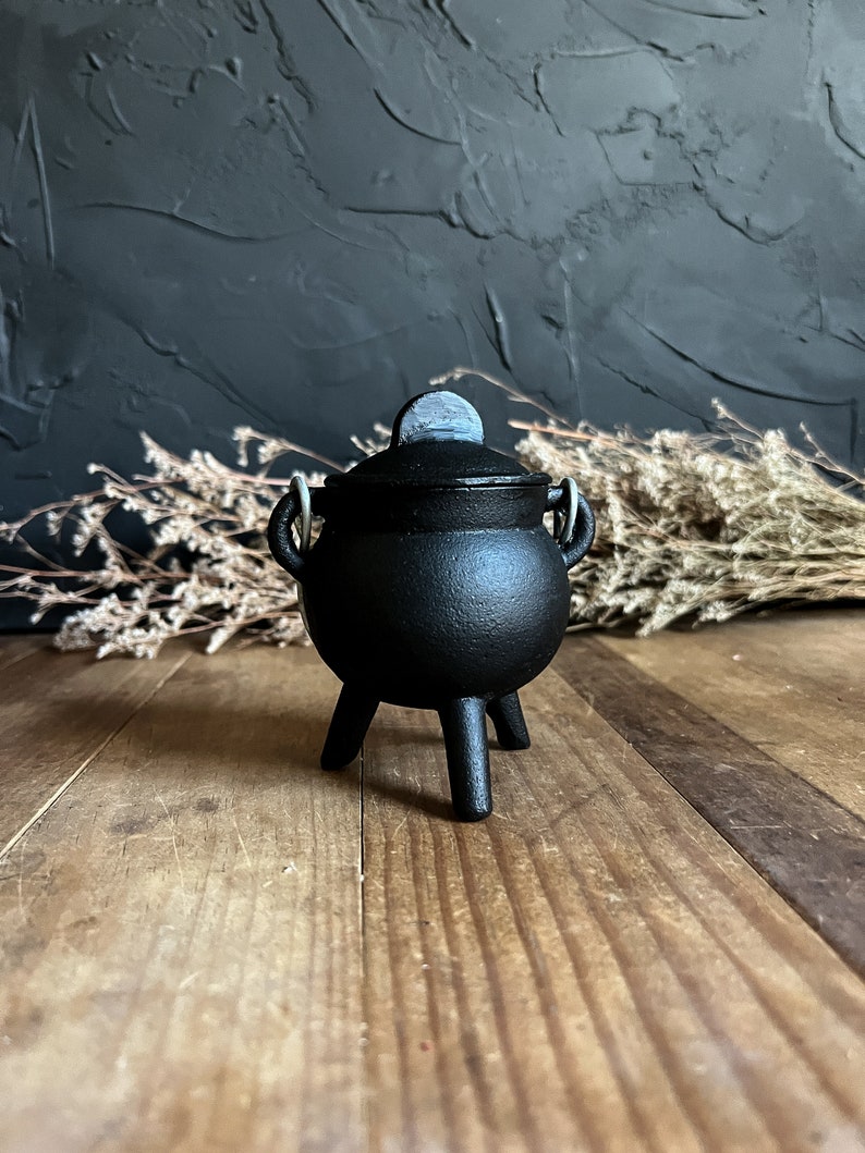 May include: A small black cast iron cauldron with a lid and three legs. The cauldron is sitting on a wooden surface.