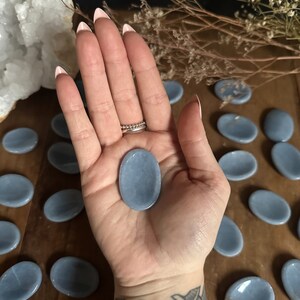 May include: A collection of smooth, light blue oval-shaped stones. The stones are arranged in a semi-circle on a wooden surface. One stone is being held in a hand.