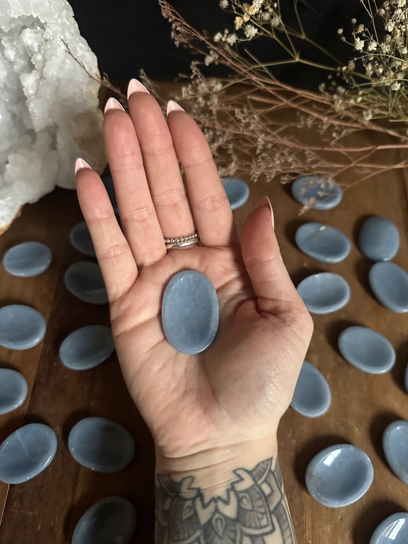 May include: A collection of smooth, light blue oval-shaped stones. The stones are arranged in a semi-circle on a wooden surface. One stone is being held in a hand.