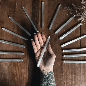 May include: A collection of clear quartz crystal wands arranged in a circular pattern on a wooden surface. A hand holds one of the wands in the centre of the circle.