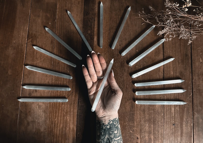 May include: A collection of clear quartz crystal wands arranged in a circular pattern on a wooden surface. A hand holds one of the wands in the centre of the circle.