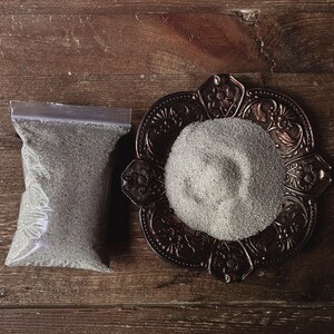 May include: A small plastic bag filled with a gray powder next to a decorative metal dish with a pile of the same gray powder.